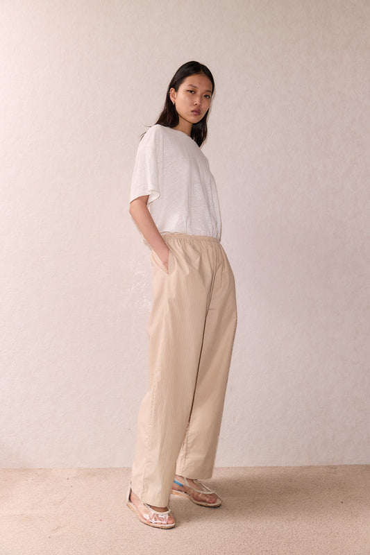 A woman with long dark hair stands against a light textured wall, wearing the ease trouser - sand stripe, a loose white t-shirt, and pink shoes. The trousers are wide-legged with an elastic waist; her hands rest in her pockets.