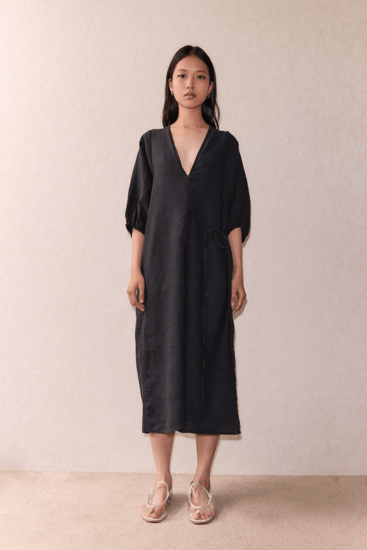 A woman faces forward against a light beige wall, wearing the Crease Bind Dress—a loose-fitting, dark wrap dress with three-quarter sleeves—and light-colored sandals.