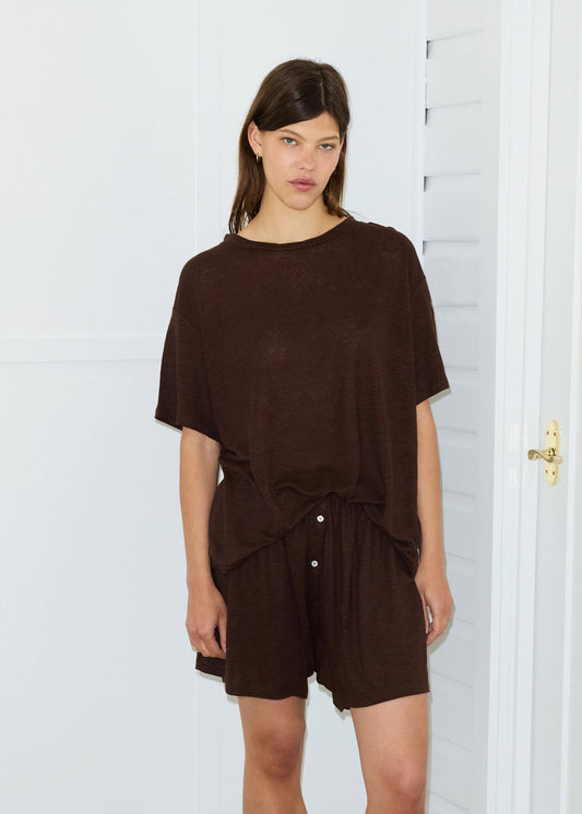 A woman with straight brown hair stands indoors in front of a white wall and door, wearing a loose-fitting dark brown Soft T-Shirt and matching shorts, looking directly at the camera with a neutral expression.