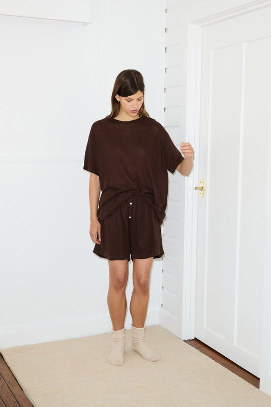 A woman stands indoors on a beige rug, wearing the Soft Short and a matching loose brown top with white buttons. She has light beige socks on and looks down with one hand slightly raised. The white room features wooden floors.