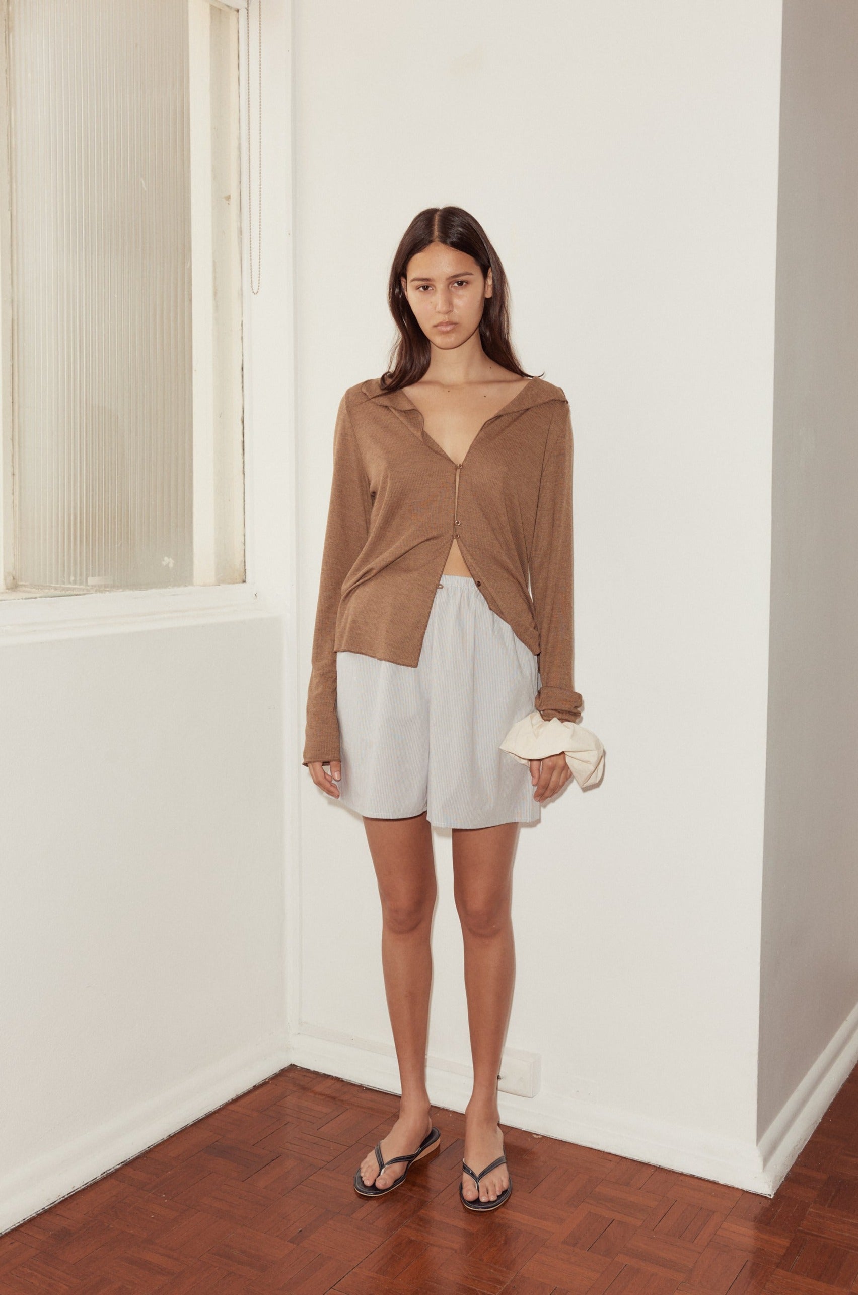 A woman stands indoors near a white wall, wearing a loose brown long-sleeve top, mid short in light gray, and black flip-flops. She holds a scrunchie in one hand and looks at the camera with a neutral expression.