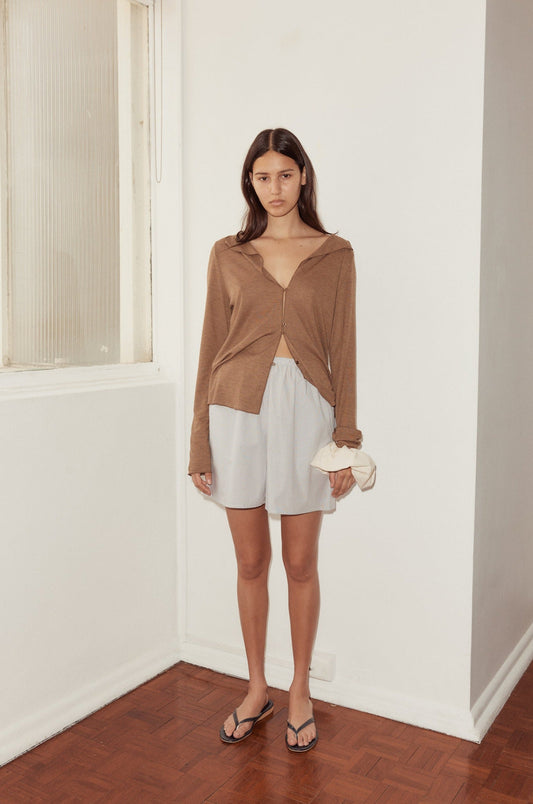 A woman stands indoors near a white wall, wearing a loose brown long-sleeve top, mid short in light gray, and black flip-flops. She holds a scrunchie in one hand and looks at the camera with a neutral expression.