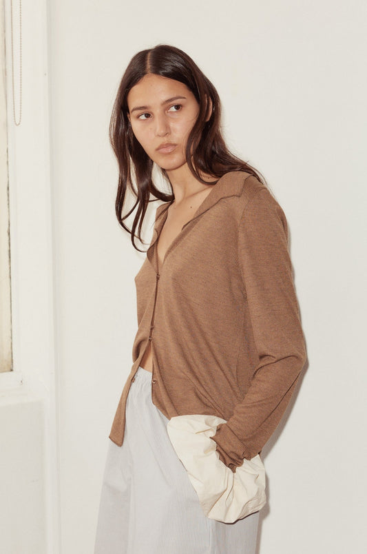 A woman with long brown hair wears the Open Long Sleeve Top, styled off one shoulder, paired with light pants. She stands against a plain white wall, looking to the side.