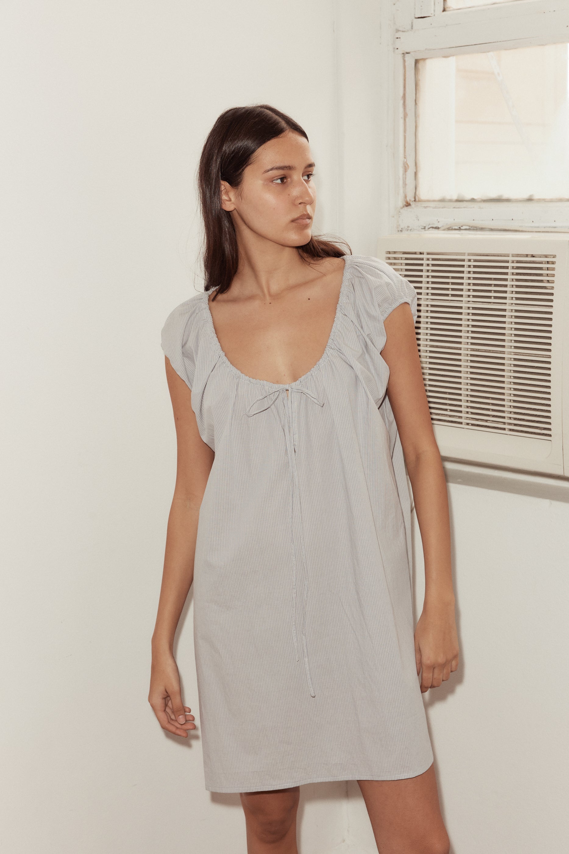 A woman with long dark hair stands indoors near an air conditioner, wearing the Capped Sleeve Dress in light gray. The dress is short-sleeved, loose-fitting, with a deep scoop neckline and tie detail at the chest. She looks to the side.