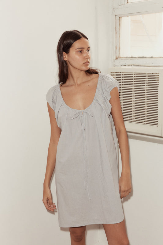 A woman with long dark hair stands indoors near an air conditioner, wearing the Capped Sleeve Dress in light gray. The dress is short-sleeved, loose-fitting, with a deep scoop neckline and tie detail at the chest. She looks to the side.