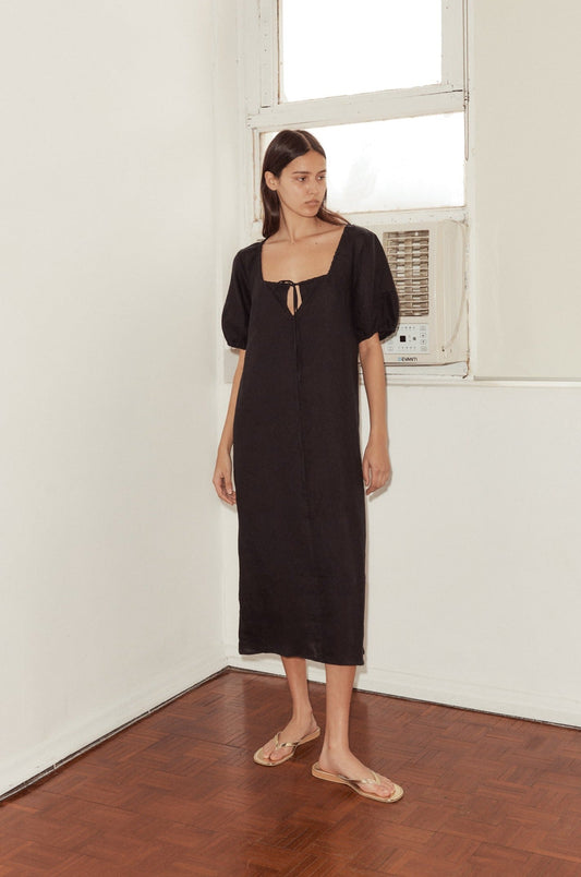 A woman wears the Curved Seam Midi Dress—black, loose-fitting, with short sleeves and a small front cutout—and tan sandals while standing indoors near a window and air conditioner, looking to the side.
