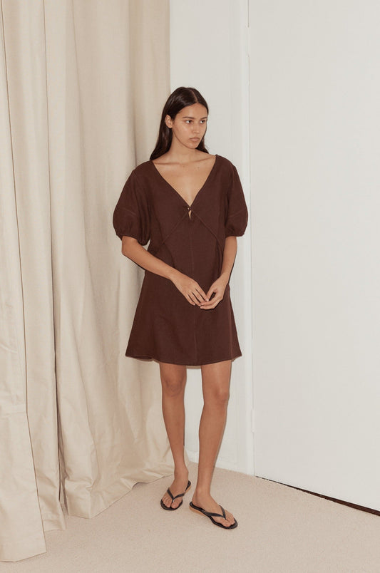 A woman with long dark hair stands indoors by a beige curtain and white door, wearing the Tie Seamed Short Dress in brown with puffed sleeves and a deep V-neckline, paired with black flip-flops.