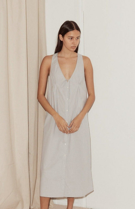 A woman with long, straight hair stands indoors in the Tuck Tie Dress, a sleeveless, light-colored button-down style with a V-neckline. She faces slightly to the side with a neutral expression; a beige curtain is behind her.