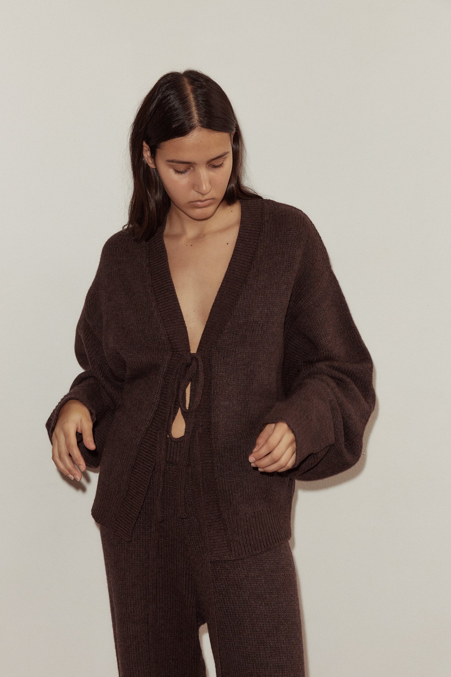 Close up shot of model wearing the Two Tie Cardigan in Plum by Deiji Studios, styled with the Straight Leg Knit Pant in Plum. Cardigan features v neckline with two long knit ties at front, one at bust and one at waist.