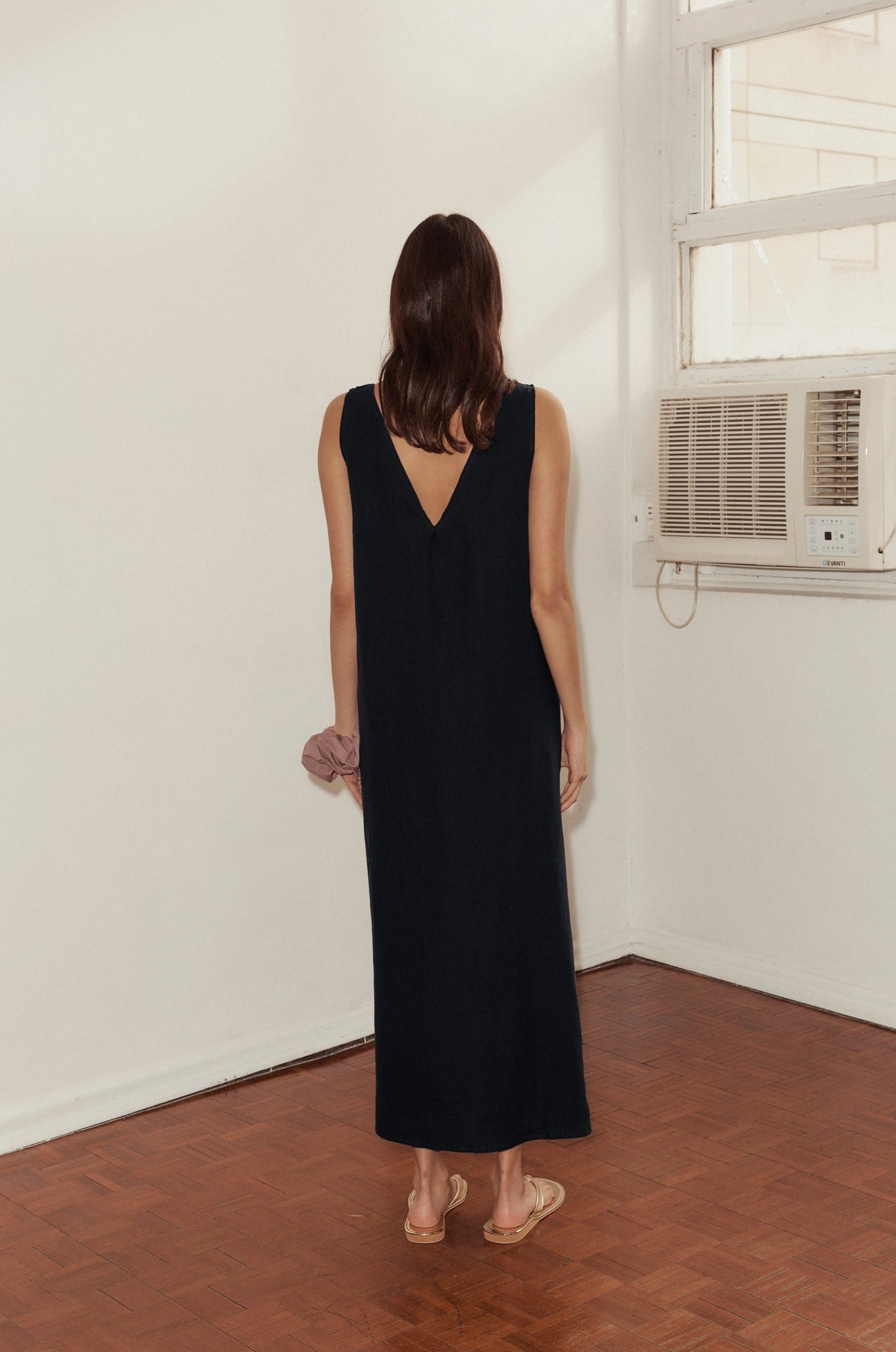 A woman with shoulder-length brown hair, wearing a sleeveless black Keyhole Dress and sandals, stands indoors on a wooden floor facing a white wall with a window and an air conditioner.