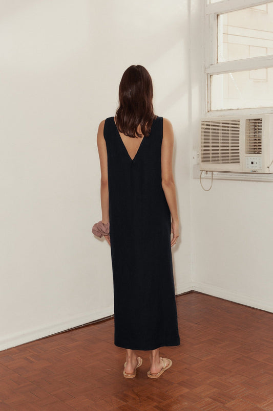 A woman with shoulder-length brown hair, wearing a sleeveless black Keyhole Dress and sandals, stands indoors on a wooden floor facing a white wall with a window and an air conditioner.