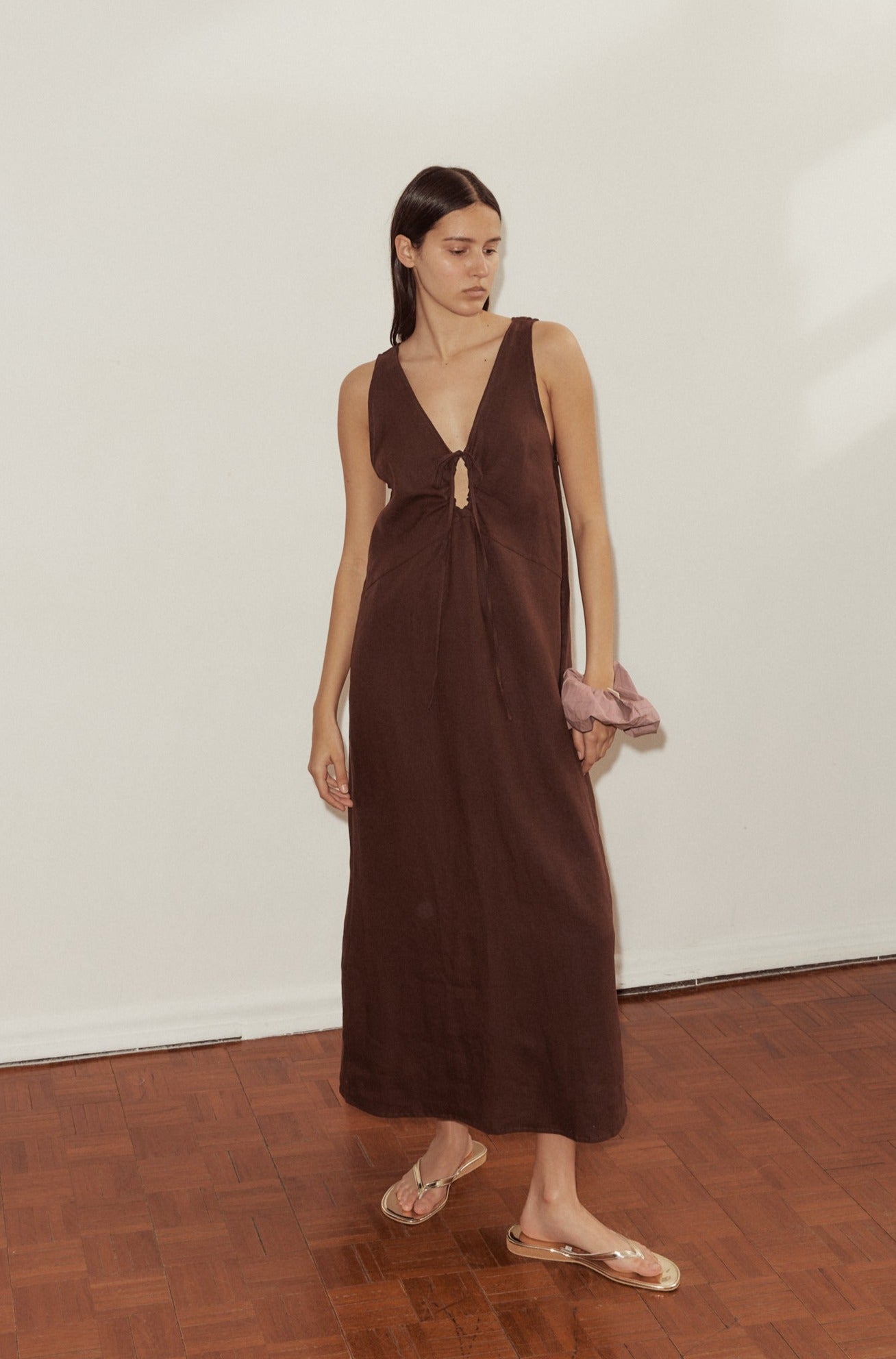 A woman stands indoors on a wooden floor, wearing the sleeveless, brown Keyhole Dress with a front tie and clear sandals. She holds a light pink scrunchie in one hand and gazes off to the side.