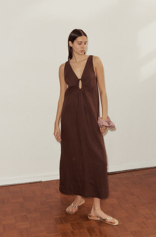 A woman stands indoors on a wooden floor, wearing the sleeveless, brown Keyhole Dress with a front tie and clear sandals. She holds a light pink scrunchie in one hand and gazes off to the side.