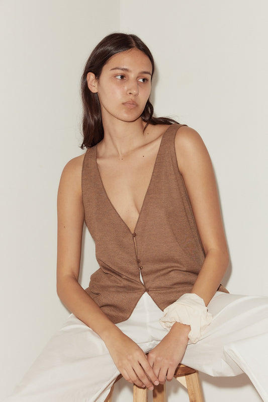 A young woman with long dark hair sits on a stool against a plain light wall, wearing the Button Up Knit Tank in brown and loose white pants. She accessorizes with a light scrunchie on her wrist and has a neutral expression.
