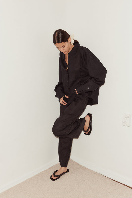 Wearing the Tack Set—a loose black button-up shirt and pants—and black sandals, a person with dark hair tied back stands in a corner with white walls and beige carpet, looking down.