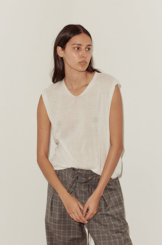 A young woman with long dark hair wears a Loose Knitted Vest and plaid pants, standing against a plain off-white wall and looking slightly to her left with a neutral expression.