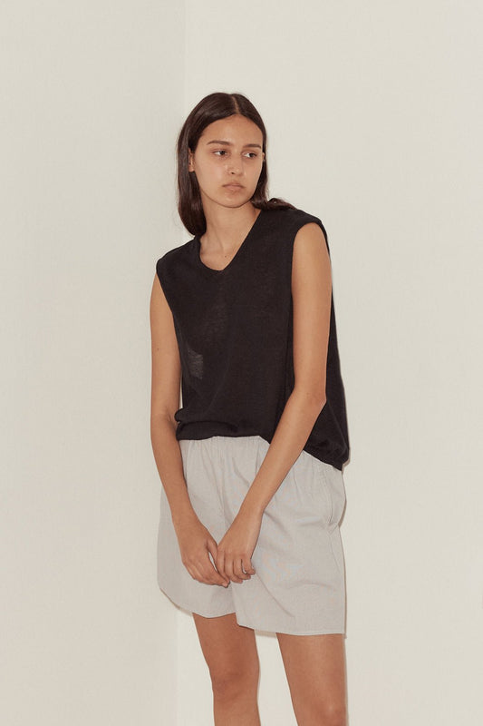 A young woman with long dark hair stands indoors against a beige wall, wearing the Loose Knitted Vest in black and light gray shorts. She has a neutral expression and rests her left hand in her pocket.