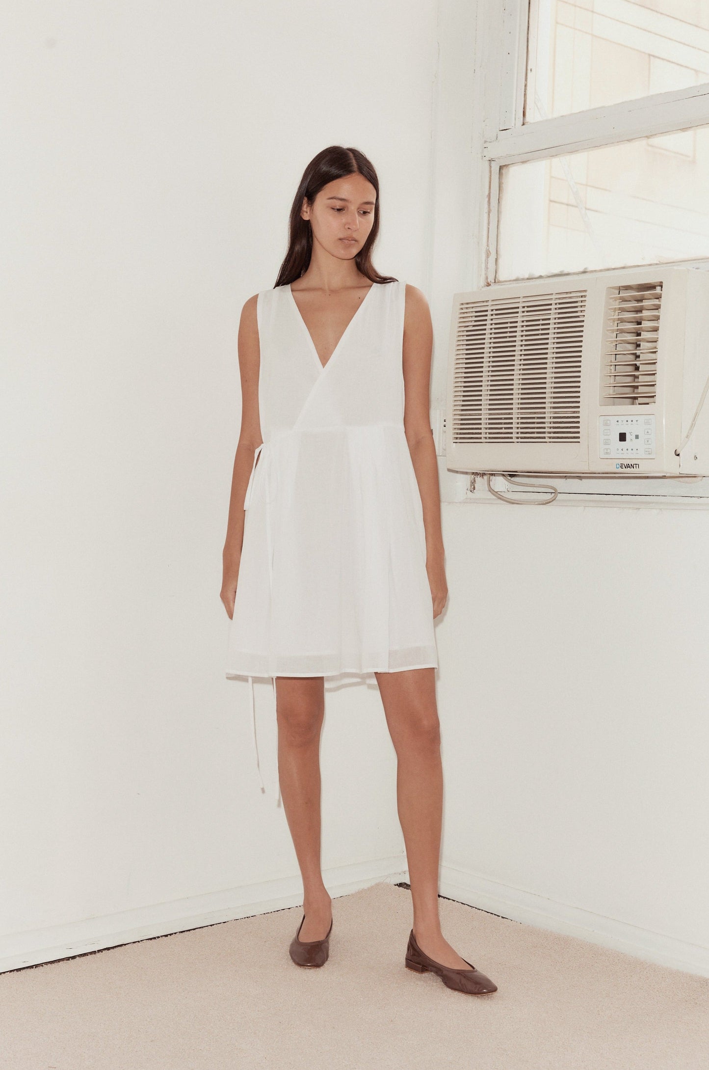 A woman wearing the Wrap Tie Dress and brown flats stands indoors beside a window with a wall-mounted air conditioner, in a room with white walls and beige carpet.