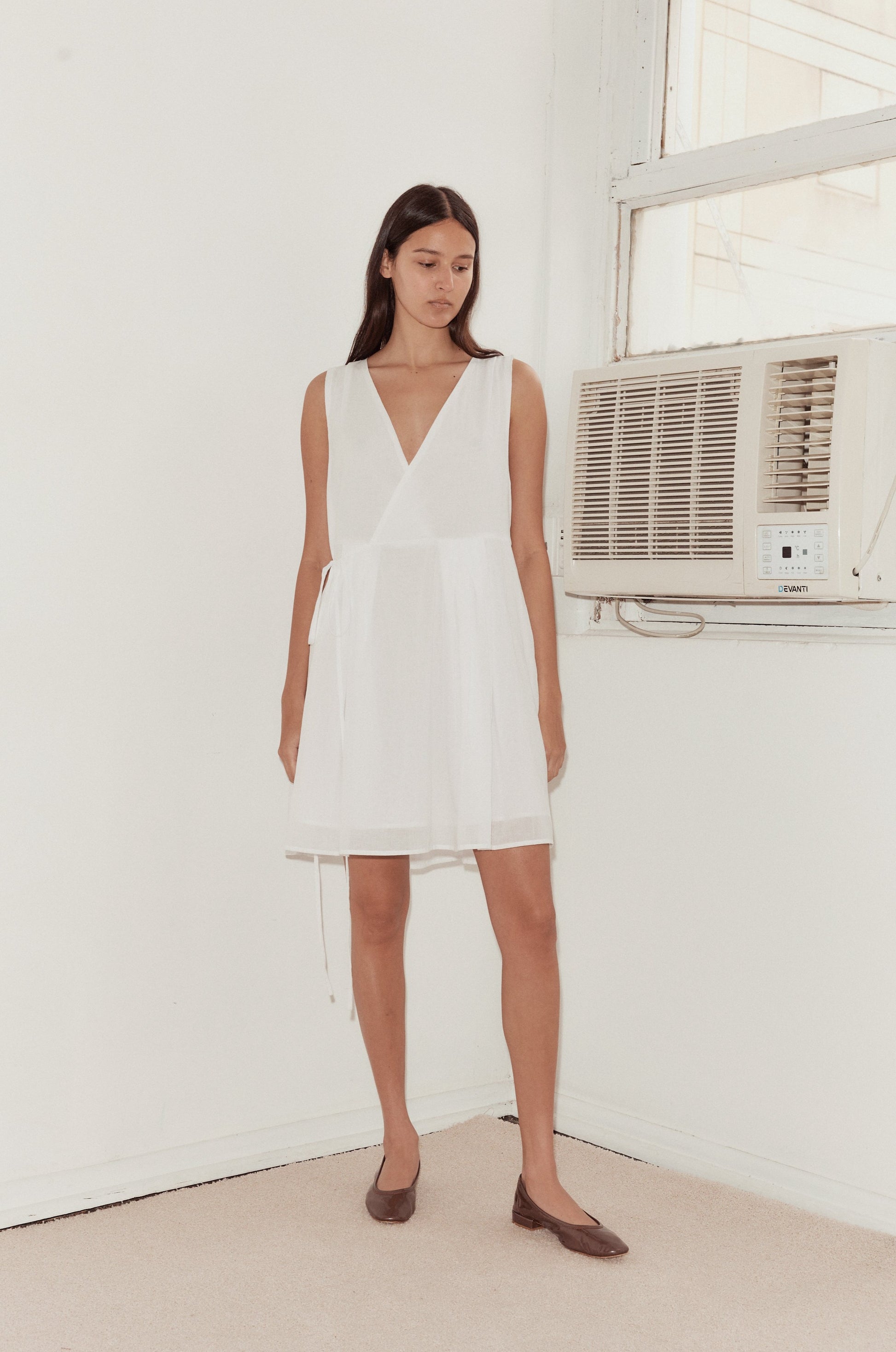 Mid shot of female model standing against a wall with a vintage airconditioning unit wearing the Wrap Tie Dress in white by Deiji Studios. A relaxed airy mini dress with v neckline, waist tie and double lined skirt.