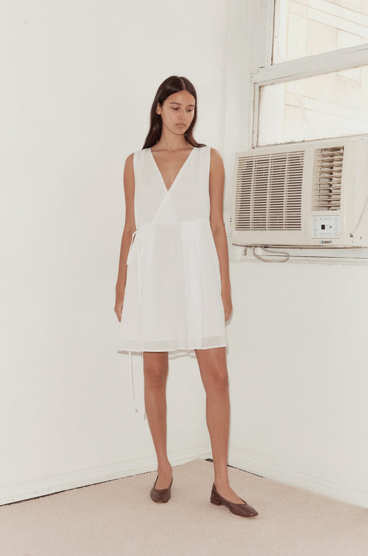 Mid shot of female model standing against a wall with a vintage airconditioning unit wearing the Wrap Tie Dress in white by Deiji Studios. A relaxed airy mini dress with v neckline, waist tie and double lined skirt.