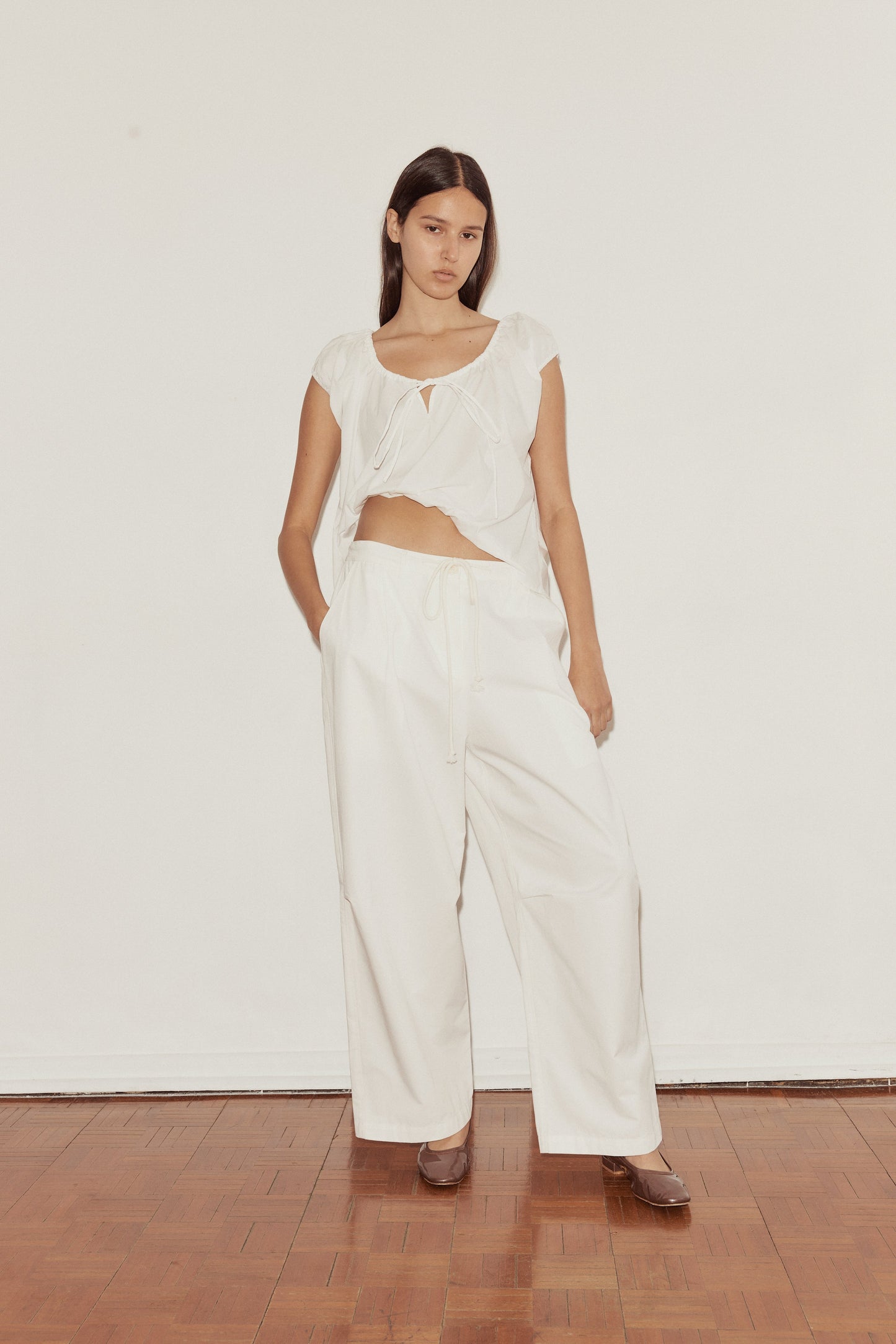 A woman stands on a wooden floor against a white wall, wearing wide-legged white Bartack Pant and a loose, short-sleeved top with an asymmetrical hem. She faces the camera with a neutral expression.