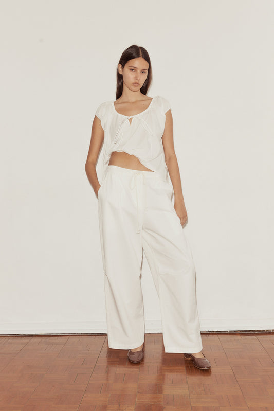 A woman stands on a wooden floor against a white wall, wearing wide-legged white Bartack Pant and a loose, short-sleeved top with an asymmetrical hem. She faces the camera with a neutral expression.