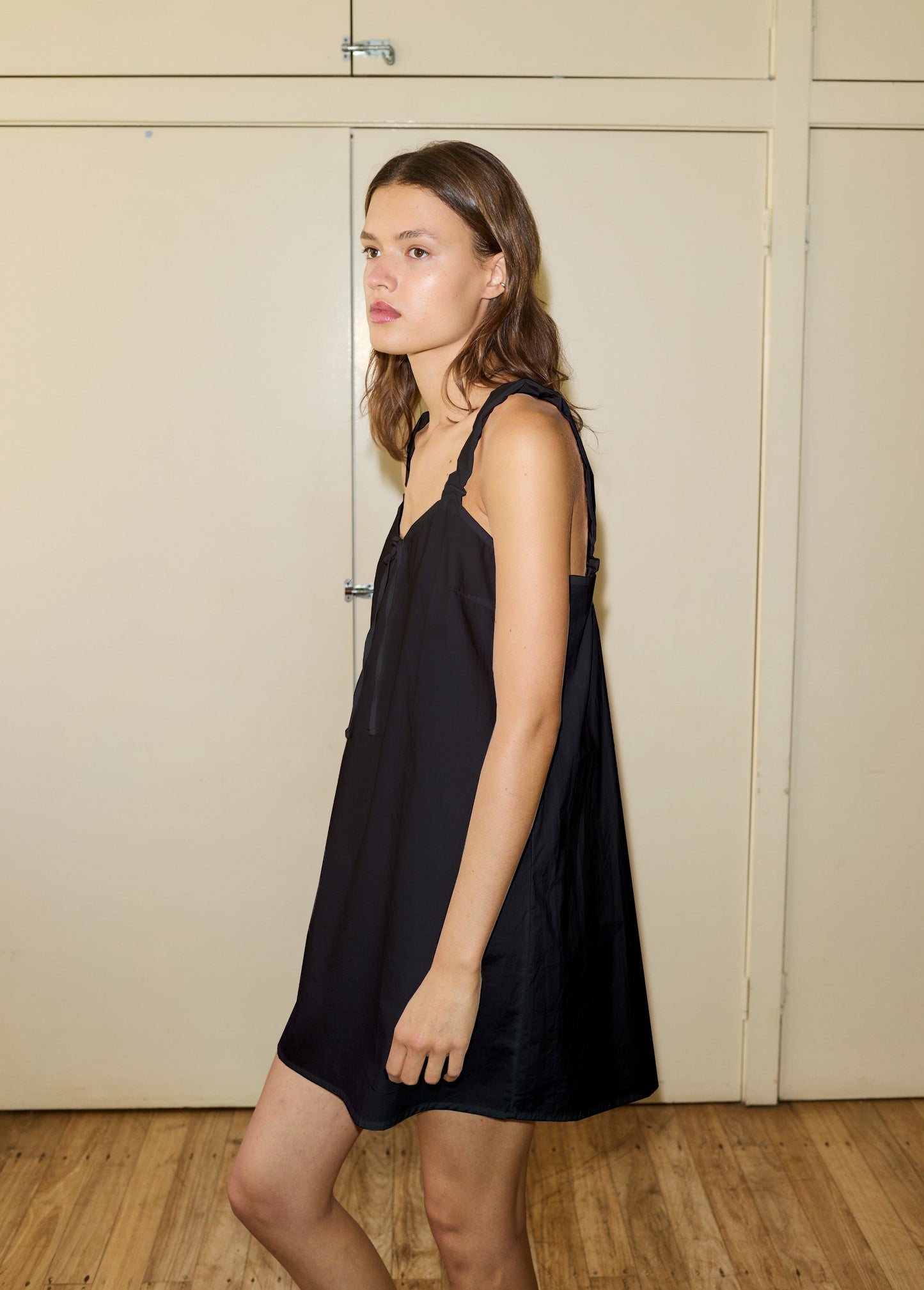 A young woman with light brown hair wears a loose, sleeveless black cotton slip and stands indoors on a wooden floor facing left, her expression neutral. The background features light beige paneled doors.