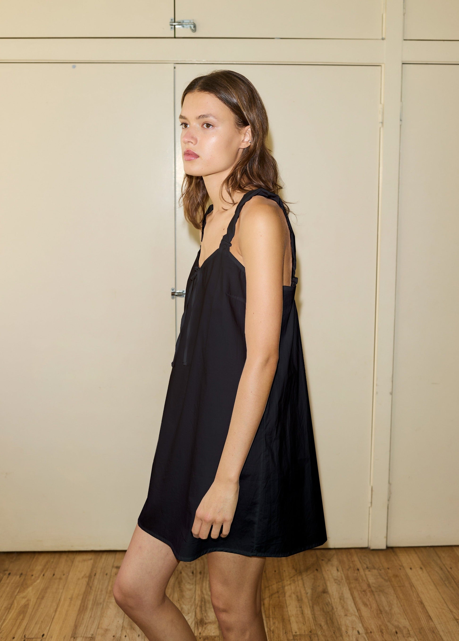A young woman with light brown hair wears a loose, sleeveless black cotton slip and stands indoors on a wooden floor facing left, her expression neutral. The background features light beige paneled doors.