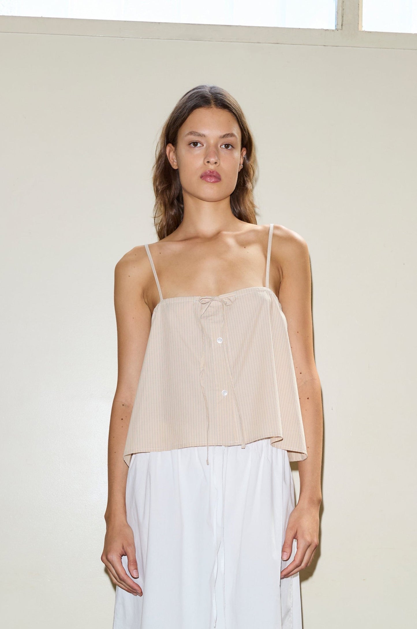 A woman with wavy brown hair stands against a beige wall, wearing the sleeveless Placket Strap Top in beige with thin straps and white bottoms, looking directly at the camera with a neutral expression.