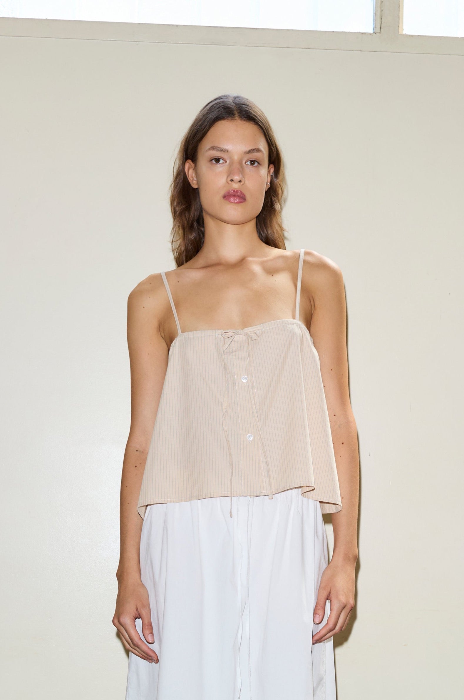 A woman with wavy brown hair stands against a beige wall, wearing the sleeveless Placket Strap Top in beige with thin straps and white bottoms, looking directly at the camera with a neutral expression.