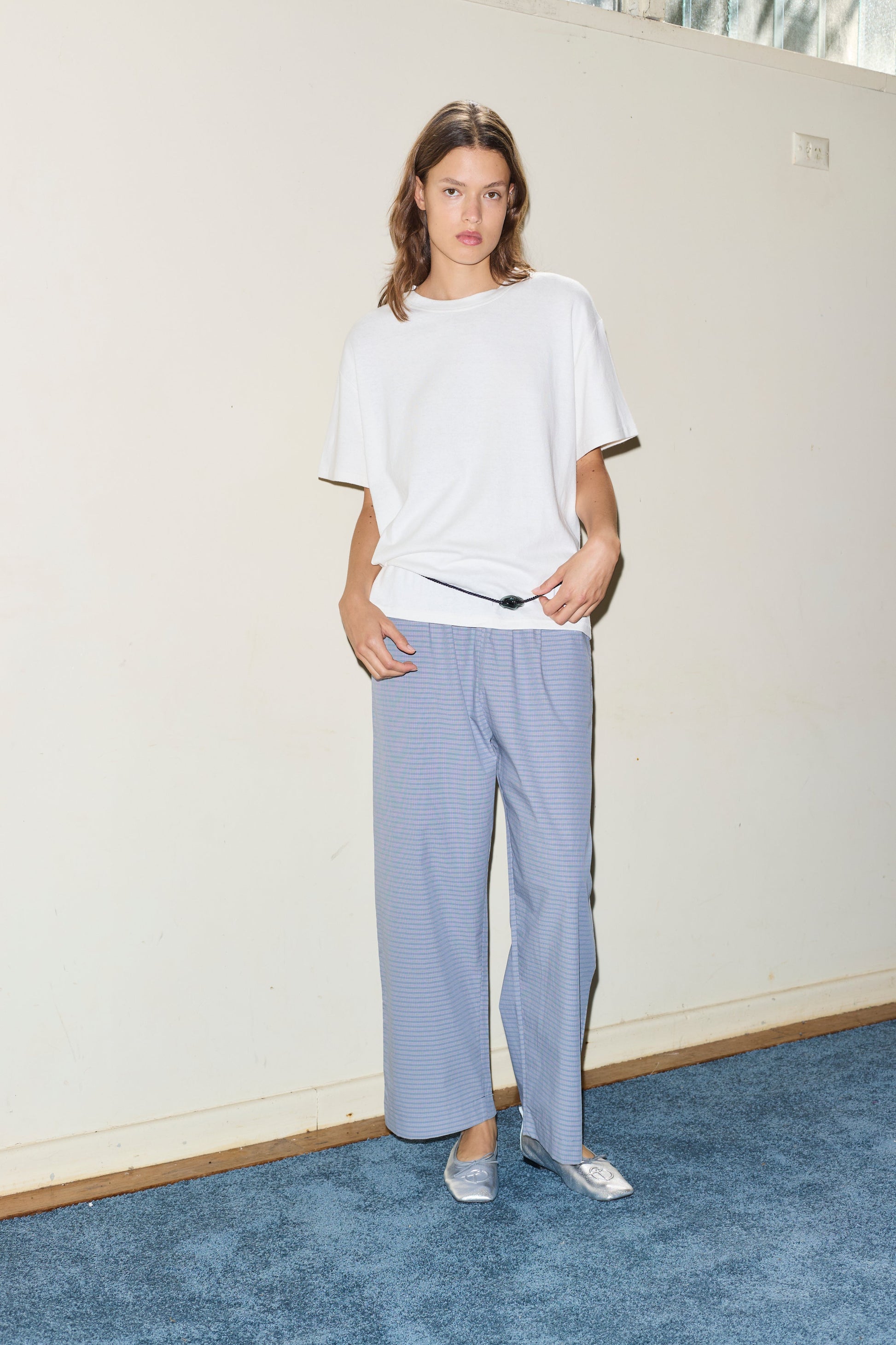 A woman with shoulder-length brown hair stands indoors on blue carpet, wearing a loose white t-shirt, silver flats, and the Ease Trouser in light blue. She has a neutral expression and one hand in her pocket.