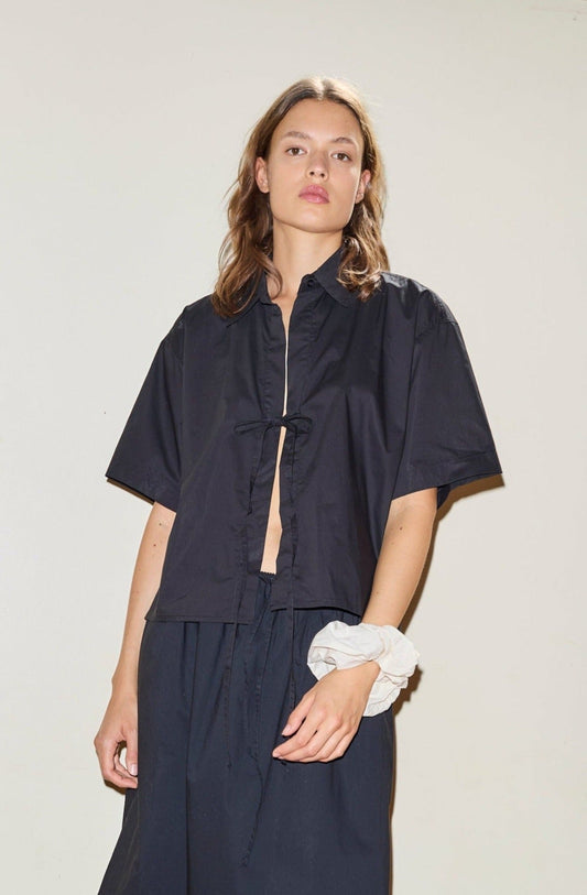 A person wearing The Tie Up Shirt, a loose-fitting, short-sleeved black shirt tied at the front with matching pants, stands against a plain light background and has a white scrunchie on their left wrist.