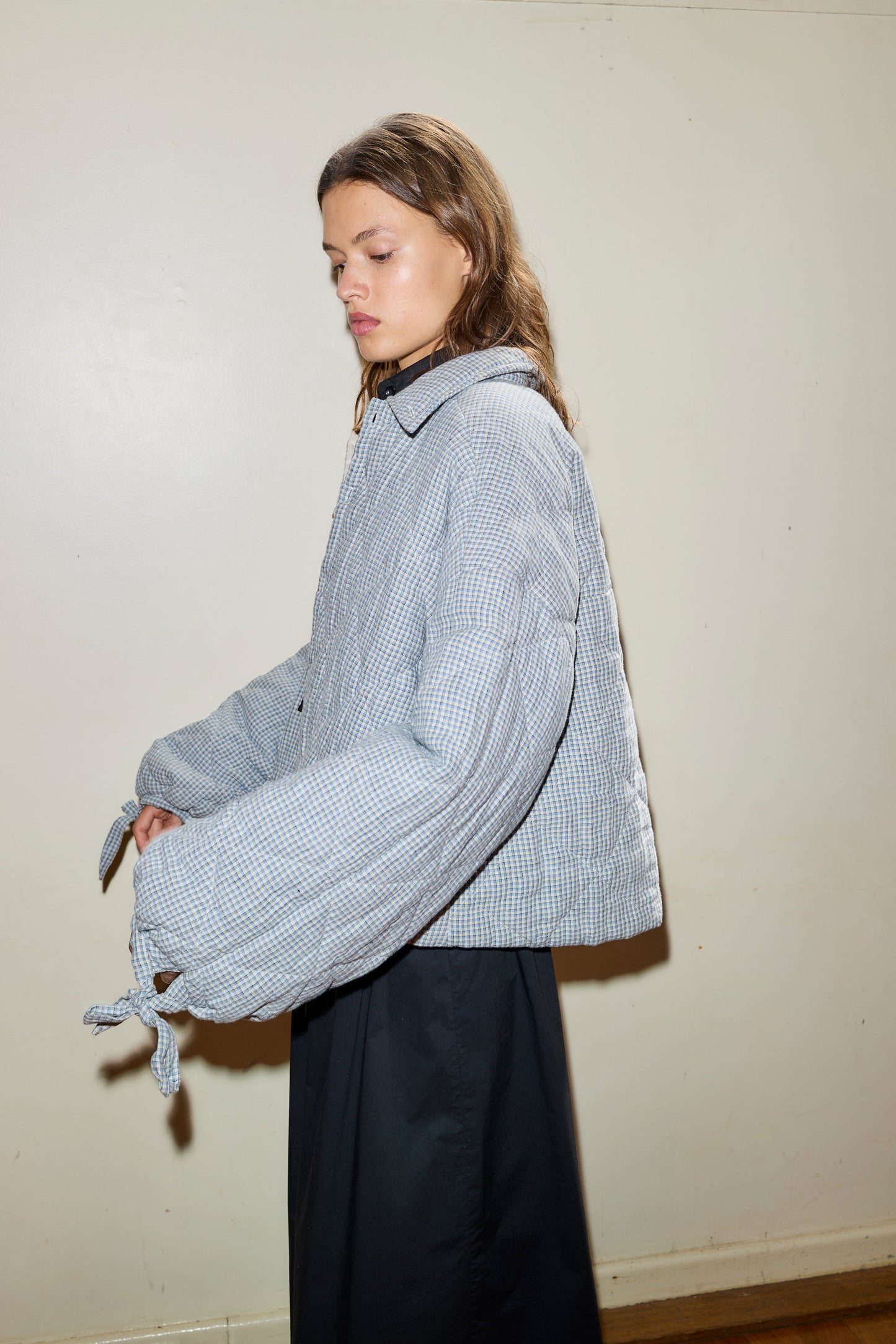 A woman with long brown hair stands against a light wall, wearing the Collared Quilt Coat—an oversized, plaid-patterned jacket with wide sleeves—paired with a dark skirt. She looks down neutrally.
