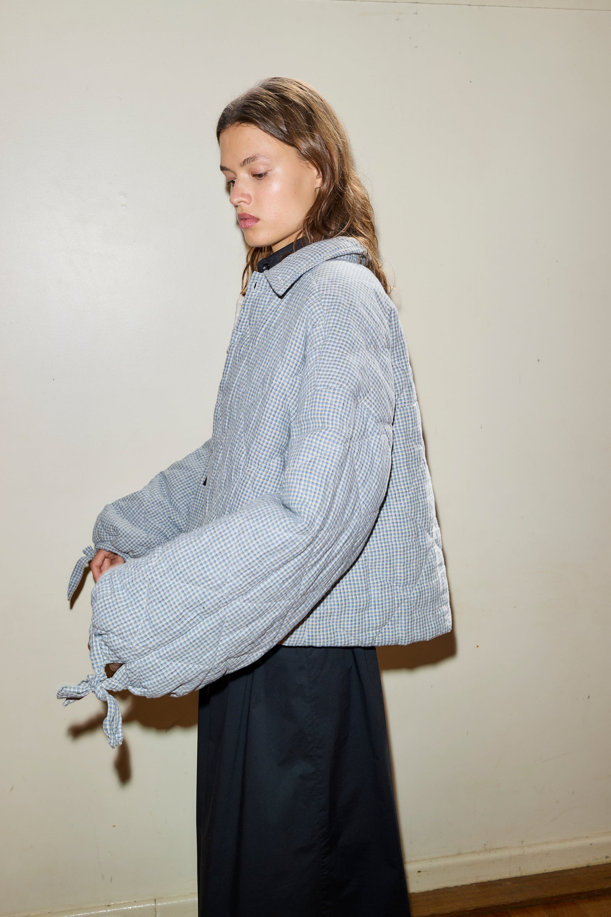 A woman with long brown hair stands against a light wall, wearing the Collared Quilt Coat—an oversized, plaid-patterned jacket with wide sleeves—paired with a dark skirt. She looks down neutrally.