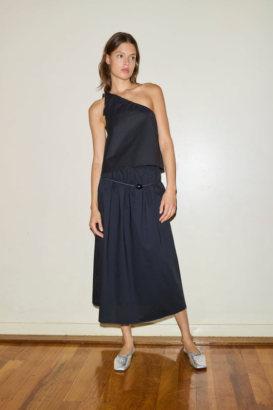 A woman with shoulder-length brown hair stands indoors against a light wall, wearing silver flats and the Mid Cotton Skirt—a black, calf-length skirt—paired with a one-shoulder black top on a wooden floor.