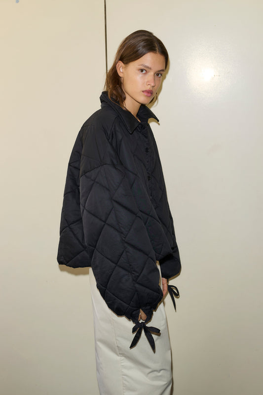 A young woman with straight brown hair wears the Collared Quilt Coat, a black quilted jacket with oversized sleeves and tie details, paired with a light skirt, standing against a plain light-colored wall.