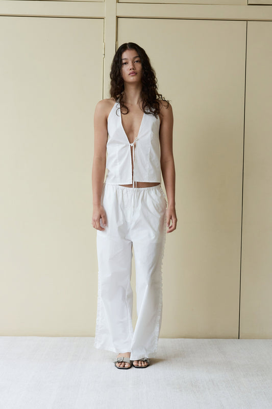 A person with long, curly hair stands indoors against a pale beige background, wearing loose white Ruffle Pants, a sleeveless white halter top with a front zipper, and black sandals.