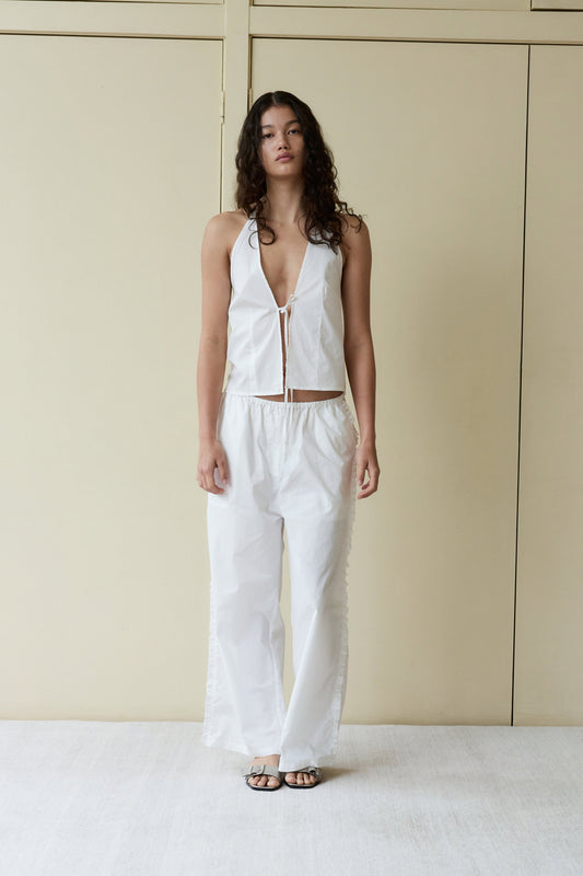 A person with long, curly hair stands indoors against a beige wall, wearing a white sleeveless halter top with a deep V-neck and matching Ruffle Pant, paired with black sandals.