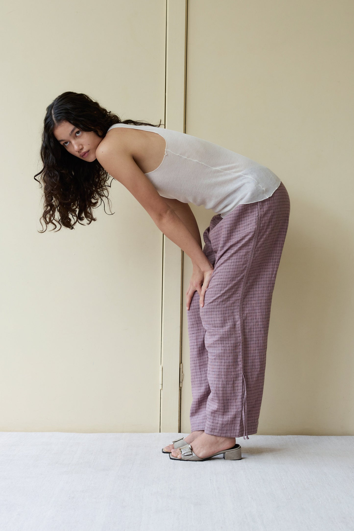 A woman with long curly hair bends forward, looking at the camera. She wears a white sleeveless top, Side Stripe Pant in mauve, and sandals, standing indoors against a beige wall and door.