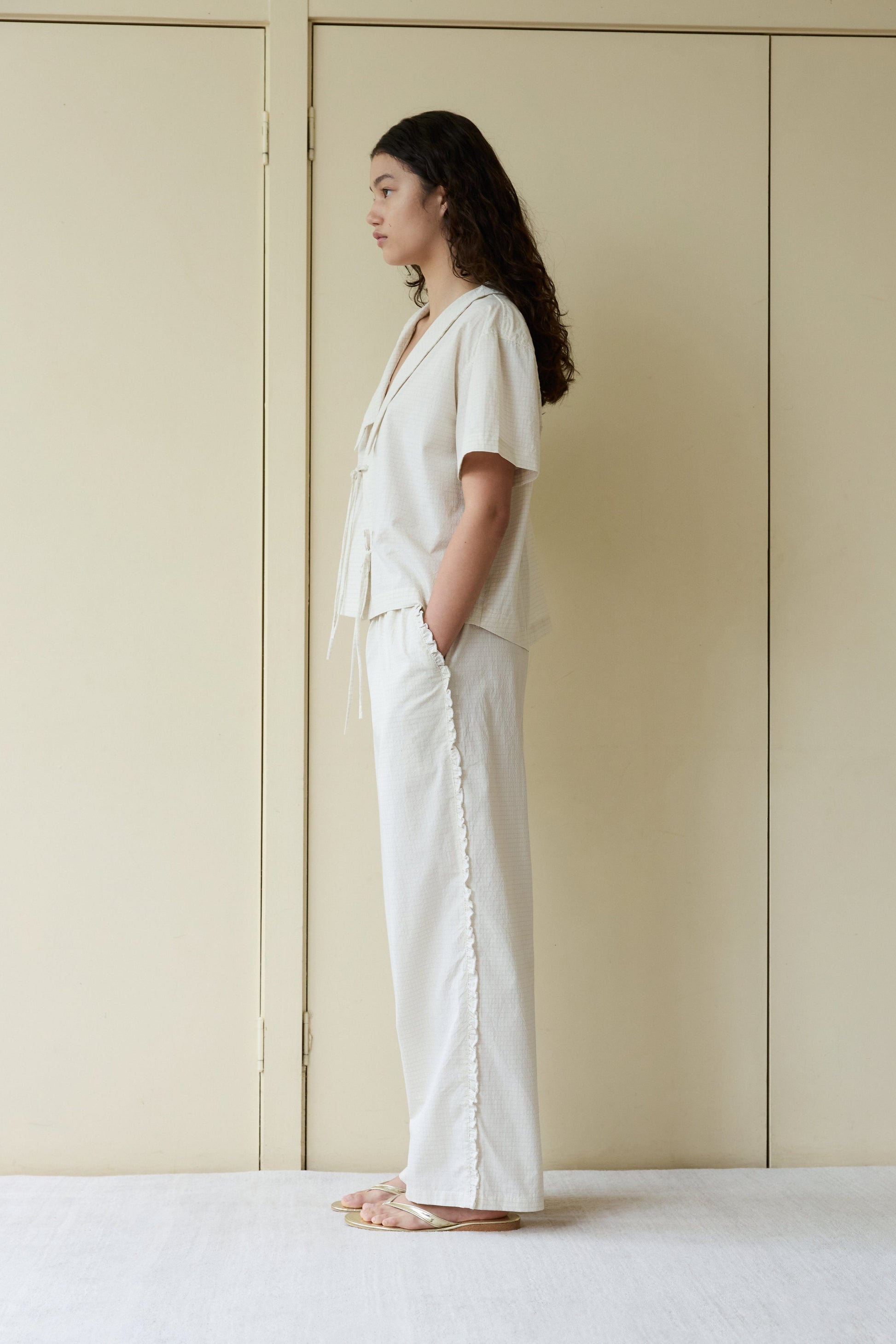 A person with long curly hair stands in profile against cream doors, wearing the Ruffle Pant—a loose, light-colored, wide-leg style—paired with a short-sleeve top and sandals. Their hands are in their pockets and their expression is neutral.