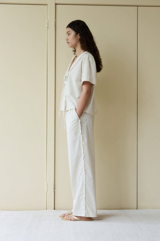 A person with long curly hair stands in profile against cream doors, wearing the Ruffle Pant—a loose, light-colored, wide-leg style—paired with a short-sleeve top and sandals. Their hands are in their pockets and their expression is neutral.