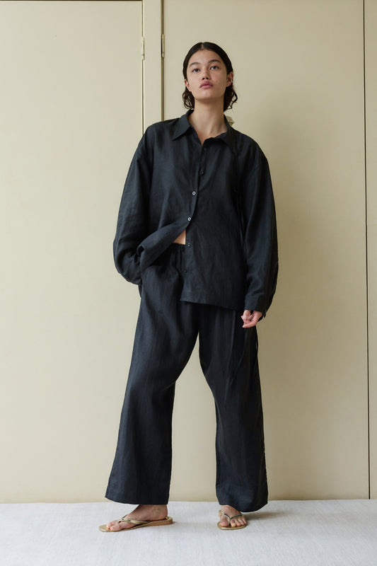 A person stands indoors against a beige wall, barefoot and confident, wearing the String Linen Set—a loose-fitting, dark button-up shirt with matching wide-leg pants.