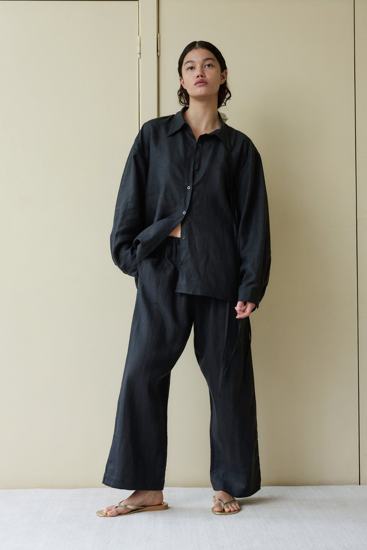 A person stands indoors against a beige wall, wearing the String Linen Set—loose, dark button-up shirt and matching wide-leg pants—with tan sandals. Their dark hair is pulled back, and they pose in a relaxed manner.