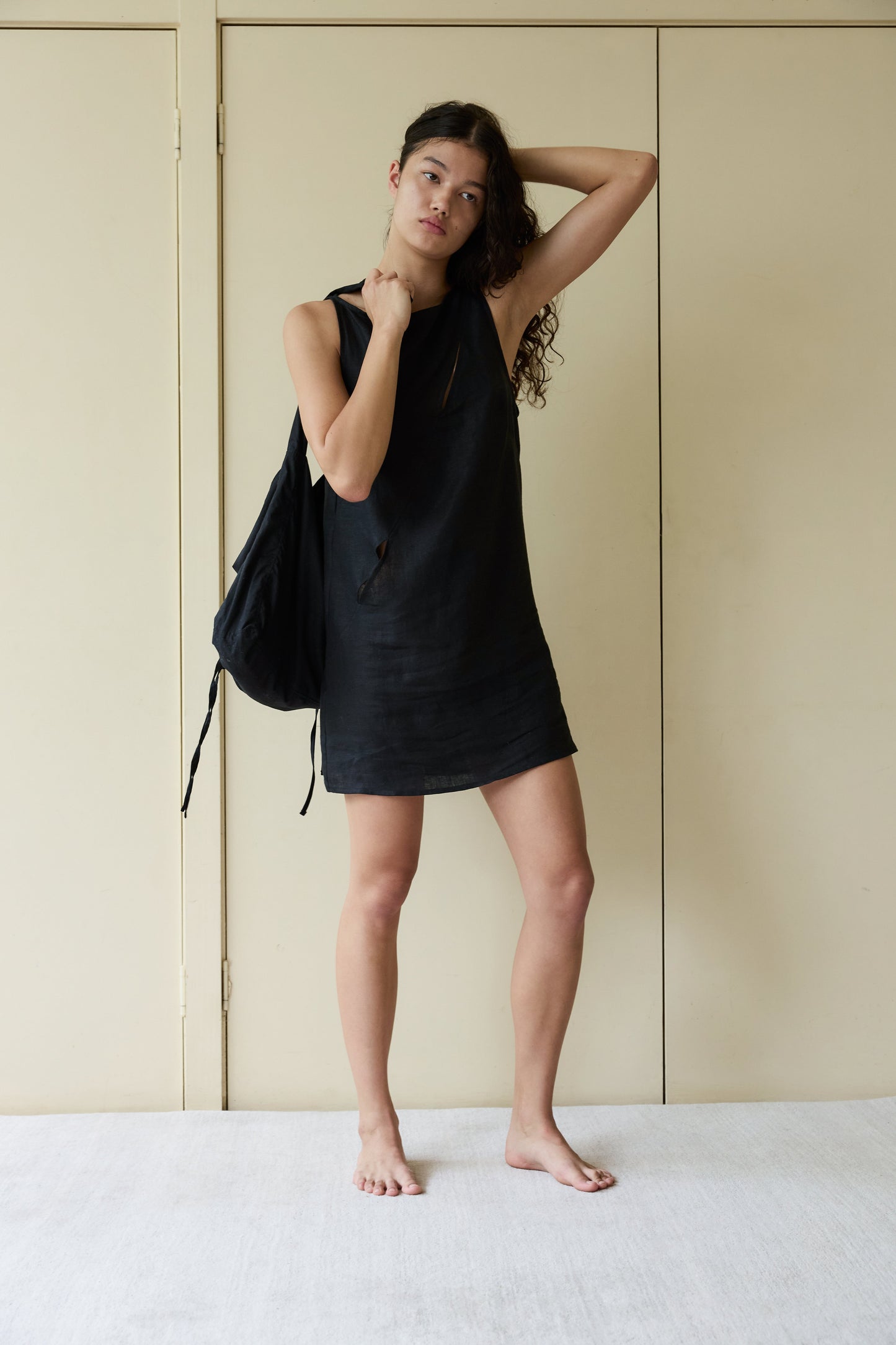 A young woman stands barefoot indoors against a beige wall, wearing the Shift Mini Dress - Black made from OEKO-TEX 100 certified stone washed French linen, holding a black backpack over her shoulder with one hand on her head.