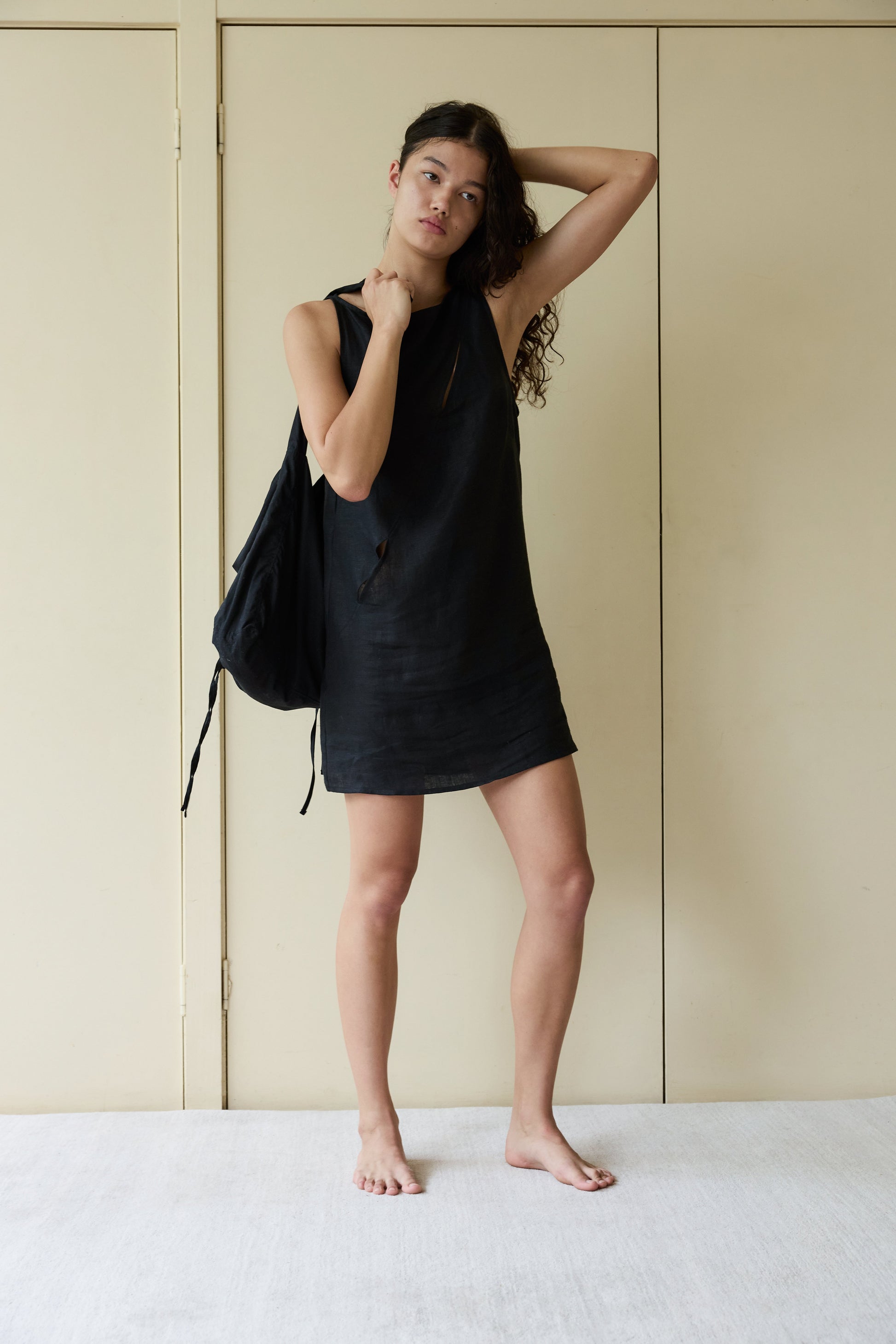 A young woman stands barefoot indoors against a beige wall, wearing the Shift Mini Dress - Black made from OEKO-TEX 100 certified stone washed French linen, holding a black backpack over her shoulder with one hand on her head.