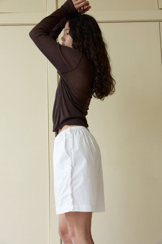A person with curly hair stands in profile against a beige background, wearing Ruffle Shorts and a long-sleeved brown top, with arms raised above their head.