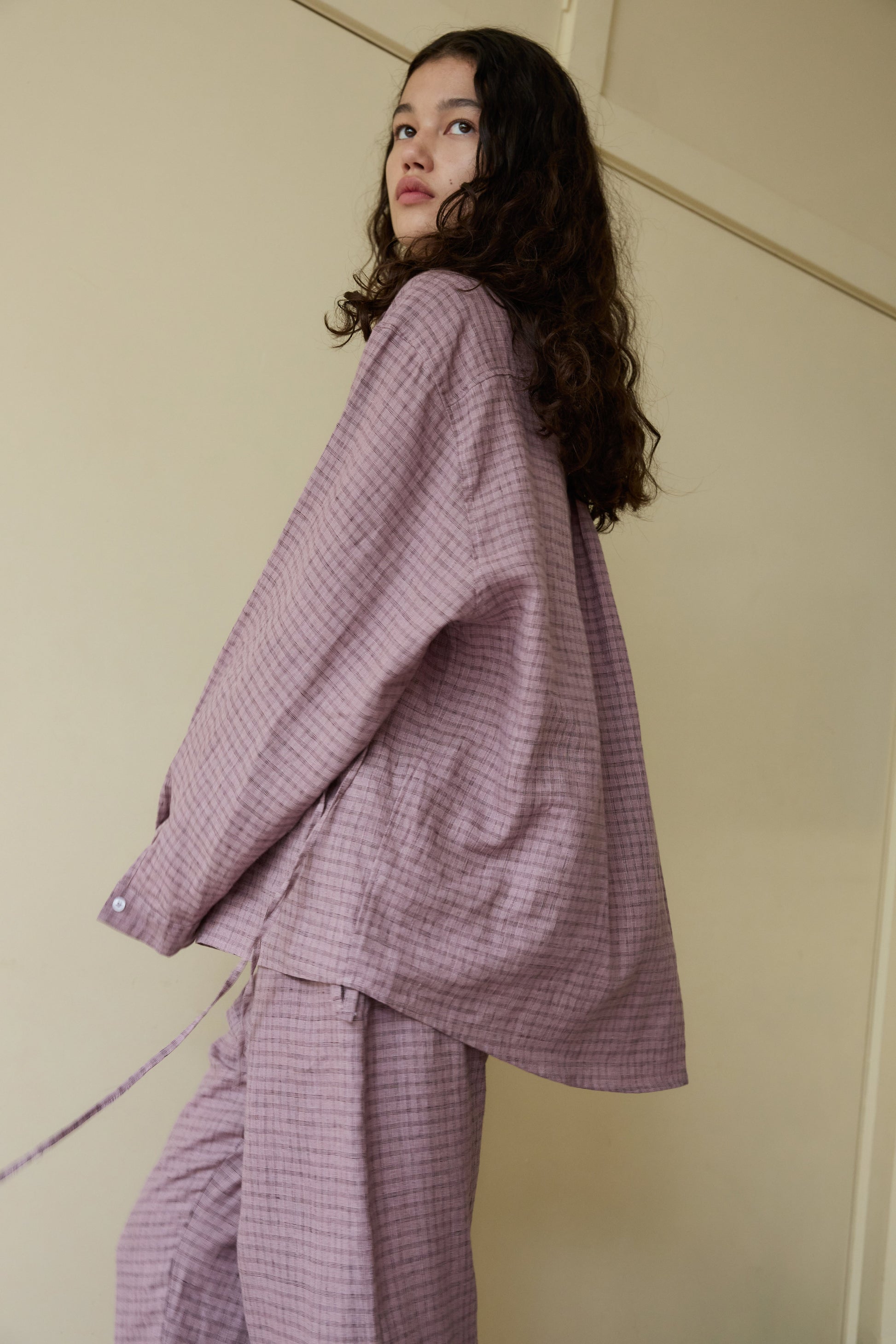 A woman with long curly hair, dressed in the loose-fitting, textured light purple String Linen Set, stands sideways and gazes upward against a cream-colored wall.