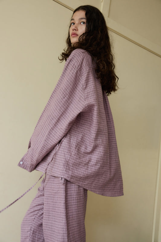 A woman with long curly hair, dressed in the loose-fitting, textured light purple String Linen Set, stands sideways and gazes upward against a cream-colored wall.
