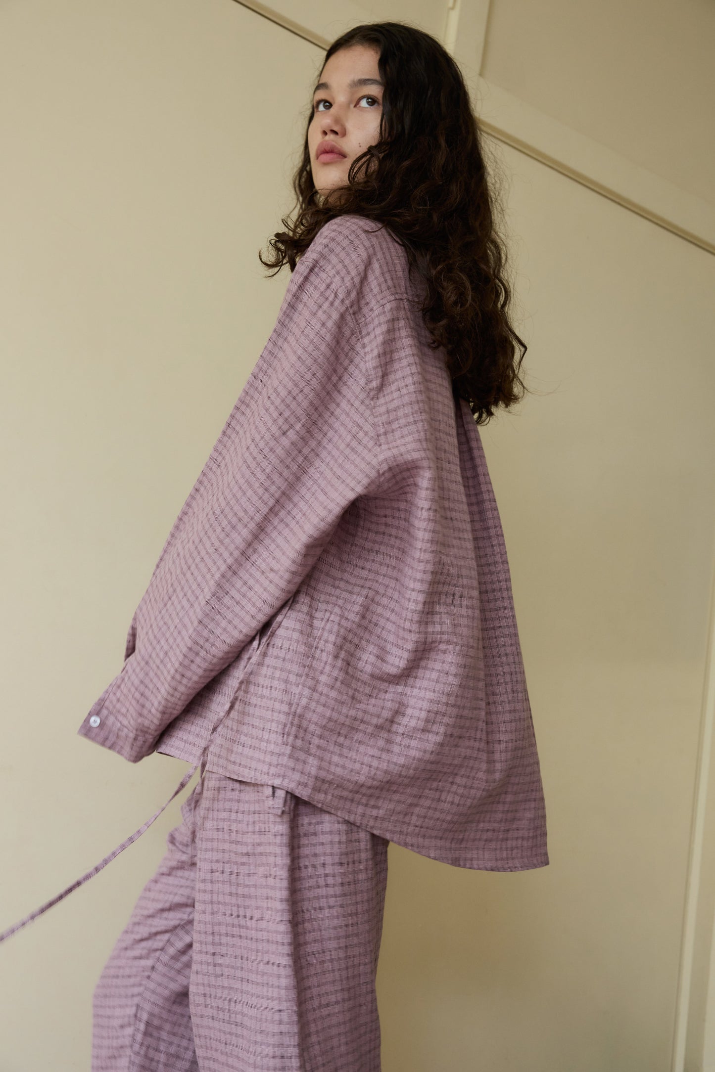 A person with long curly hair wears the String Linen Set—an oversized, textured mauve button-up shirt with matching pants—while standing indoors against plain beige walls, looking to the side.