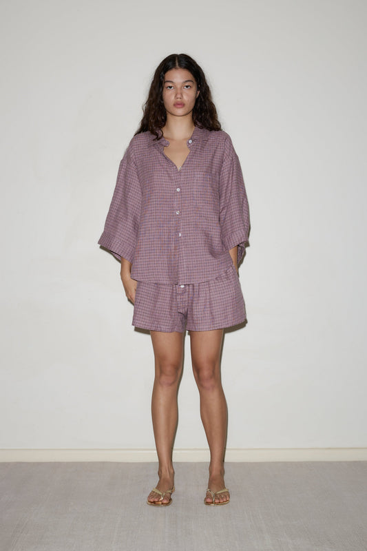 A woman with long dark hair stands against a plain light wall wearing the 03 set: a loose, long-sleeved, button-up shirt and matching purple-patterned shorts, paired with flat sandals.